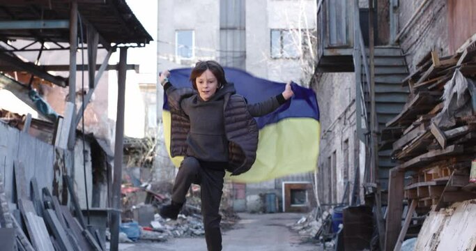 Little Ukrainian boy running and jumping on street in ruined town with national blue and yellow flag. Small kid playing outside. Destroyed city. War in Ukraine, conflict, Russian invasion, childhood