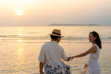 Happy Asian family couple holding hand and walking together on tropical beach at summer sunset on...