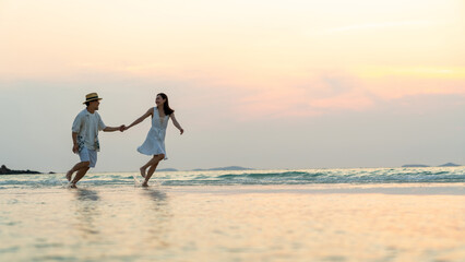 Fototapeta premium Happy Asian family couple walking and playing in sea water together on tropical beach at summer sunset on beach vacation. Husband and wife relax and enjoy outdoor lifestyle on holiday travel vacation