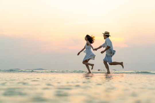 Happy Asian family couple walking and playing in sea water together on tropical beach at summer sunset on beach vacation. Husband and wife relax and enjoy outdoor lifestyle on holiday travel vacation
