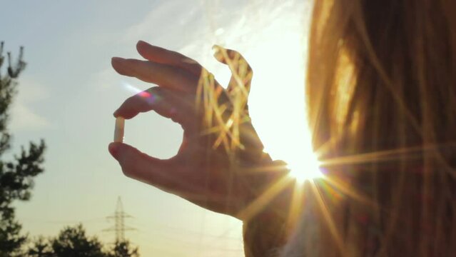 Closeup of hand holding pill tablet capsule in front of sunset yellow sun, medication, outdoor, static