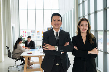 business men and women Attractive Confident Asian Standing with his arms crossed in the office with a group of colleagues working on their laptops behind the scenes.