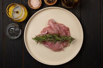 fresh raw rabbit meat on a plate with a sprig of rosemary, next to the plate spices for marinade - pink salt, a mixture of allspice, honey, olive oil, flat lay, open space