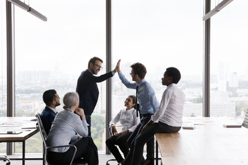 Cheerful male office friends giving high fives at business team meeting, celebrating work result,...