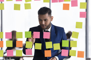 Positive engaged millennial business leader man writing notes on colorful stickers, working on team tasks management, scheduling, planning project strategy. Corporate coach preparing presentation