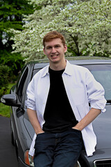 A young man next to his gray car wearing a white shirt. He has auburn red hair.