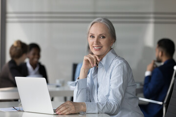 Obraz premium Happy confident mature businesswoman in open office workplace head shot portrait. Grey haired professional woman sitting at laptop computer, looking at camera, smiling. Business, leadership concept