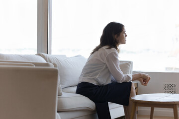 Sad depressed lonely young woman sitting on couch at home, looking at window in deep shocking...