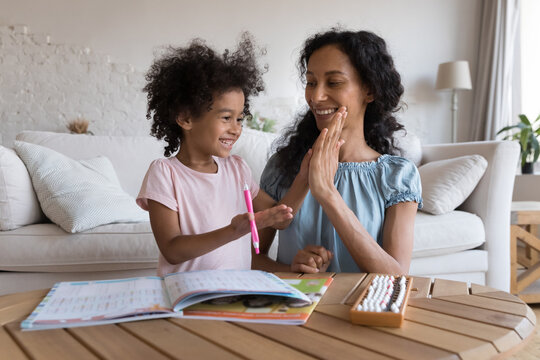 Excited African mom and cheerful kid girl clapping high five hands over school homework exercising textbooks, learning educational board games. Mother giving help, support to schoolchild daughter