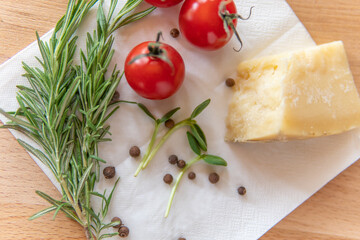 A piece of Parmesan cheese, nearby cherry tomatoes, rosemary, black peppercorns and micro greens