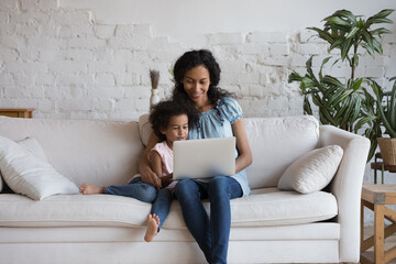 Curious adorable daughter girl and happy African mom reading online book, using learning app on laptop, watching cartoon movie, series, talking on video call, shopping. Family communication