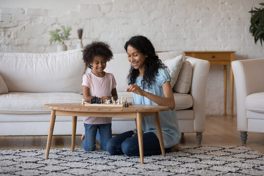 Happy African mom and daughter girl discussing game strategy at chessboard, planning next move, laughing, having fun. Daycare sitter teaching kid to play chess at home. Full length shot
