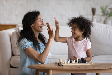 Happy excited Black mom giving high five to cute daughter over chessboard, teaching kid to play...