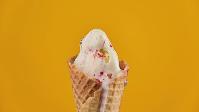 Timelapse of vanilla ice cream with strawberry topping and colorful sprinkles in waffle cone melting on yellow background. Delicious white ice cream melting. Close-up of sweet dessert. Food concept