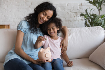 Cheerful Black mom and girl putting coin into pink piggy bank. Happy loving mother teaching little...