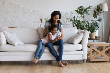 Joyful engaged Black mom teaching cute preschool daughter kid to use online learning app on mobile phone. Millennial mother and kid shopping on Internet, chatting, talking on video call in living room