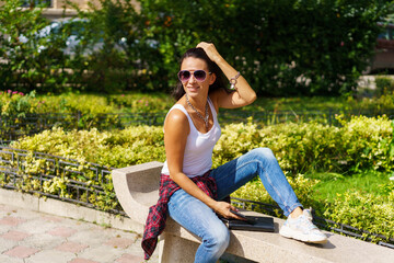 Outdoors portrait glamorous young brunette woman with glasses sitting on bench in a white t-shirt and jeans against the backdrop of a park. The photo was taken on a sunny spring or summer day.