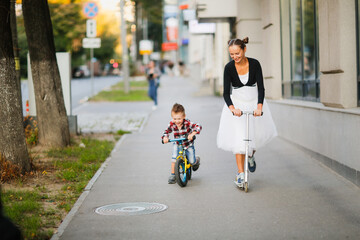 Cute young mother with her son ride on the sidewalk in the city together, child rides balance bike, mom rides scooter. Young funny mother and stylish child