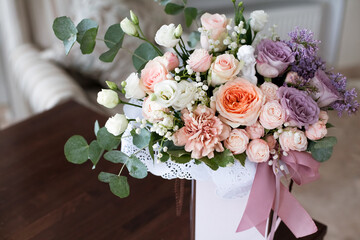 A chic festive bouquet of flowers, made of pastel-colored roses, in a decorative paper box decorated with pink ribbons, stands on a table in a bright hotel room, during the day.