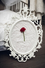 The original composition of a red rose on a white paper background in a vintage frame in a baroque style, stands on a dark wooden floor, in a bright bedroom near the bed, in the morning.