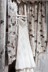 Bride morning. A stylish white wedding dress hangs on a wooden hanger on the wall of a vintage hotel with original patterned curtains.