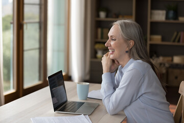 Cheerful happy mature business lady looking at window away from home workplace, smiling, laughing, sitting at table, laptop, papers, enjoying job success, online work chat