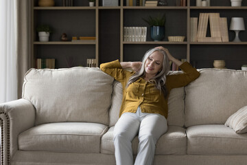 Happy pensive grey haired mature woman sitting on comfortable couch in living room, leaning on back, touching head, massaging neck, relaxing, thinking, dreaming, smiling. Home leisure concept
