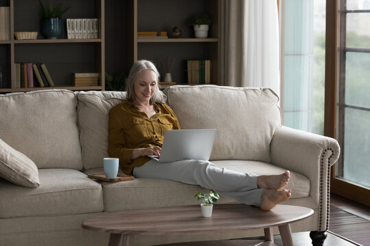 Positive mature retired grey haired woman enjoying leisure at home, resting on sofa in living room, drinking morning coffee, browsing Internet, shopping, working at laptop computer