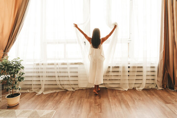 Young woman stands by large window in silk white gown after waking up at home