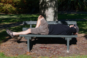 The dappled light of a tree provides shade for an older man lying on a park bench on a sunny day.