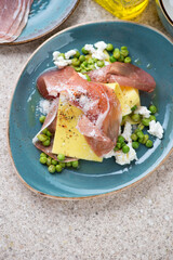 Turquoise plate with pea, ricotta and prosciutto open lasagna, flat lay on a beige marble background, vertical shot