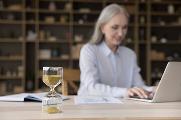 Sand glass close up on work table of senior business woman using laptop in blurred background. Mature professional keeping schedule, completing project tasks before deadline. Time management concept