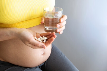 Woman in yellow top with pregnant belly hold glass of water and pills in hands. Baby expectation, health care and prenatal vitamins on pregnancy time. Illness and medicine while pregnancy.
