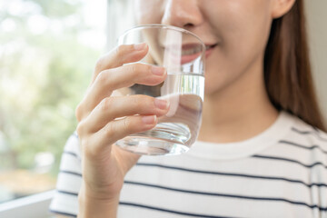 Happy beautiful, asian young woman, girl drinking, sip fresh glass of water for hydration of body, holding transparent glass in her hand, thirsty at home. Health care, healthy lifestyle concept.