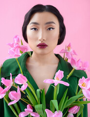Obraz na płótnie Canvas Close-up portrait of a beautiful and young Asian girl with black hair in a green cloak which is decorated with fresh pink tulips on a pink background