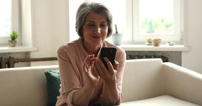 Happy smiling middle aged Hispanic woman on pension rest on couch with smartphone scroll web pages swipe photos on dating website surf news at social media. Beautiful aged lady using phone application