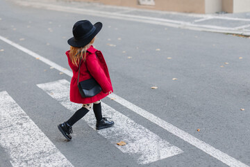 Happy little girl in a burgundy dress and hat walks along the waterfront near the pedestrian crossing in the city.