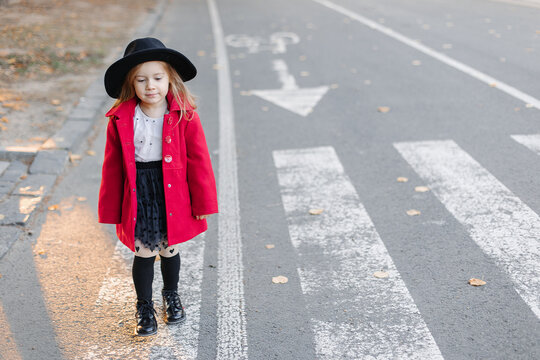 Happy little girl in a burgundy dress and hat stands at a pedestrian crossing in the city.