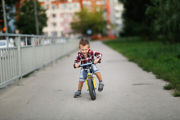cute blond european boy rides balance bike on the road in city park, child rides in city in summer, lifestyle and summer vacation in town