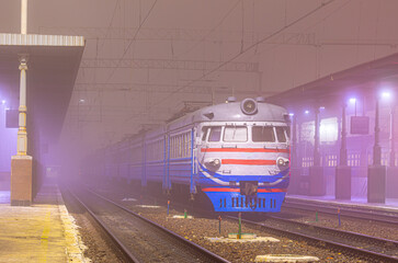 A multi-car passenger electric train is located at the platform of the railway station, waiting for departure. Foggy winter weather. Evening lighting.