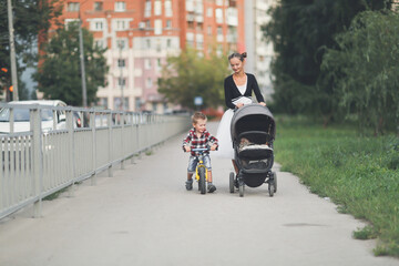 A mother with a stroller with a baby and a son on a balance bike walks around the city, a mother with siblings. child rides a balance bike