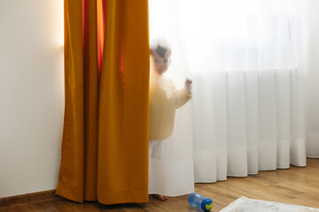 A little boy in a yellow jacket hides from his mother behind a curtain in the nursery.