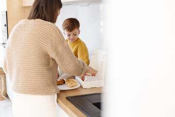 Mom is standing next to a little boy in a yellow jacket sitting on the kitchen counter next to Turkish sweets.