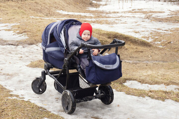 Small child in red cap in double blue pram looks at the camera and smiles. Cloudy, early spring, melting snow, last year's grass