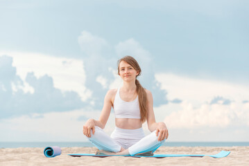 Fototapeta na wymiar Portrait of young caucasian woman sitting on yoga mat on sea background. Practice yoga outdoors on the seashore.