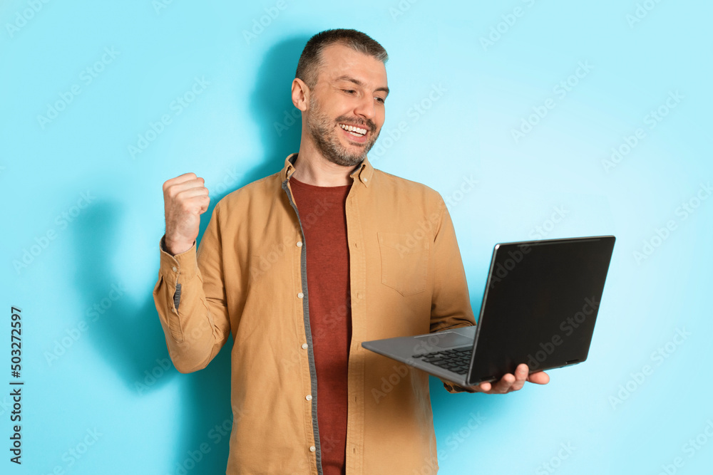 Poster joyful man using laptop shaking fists on blue background