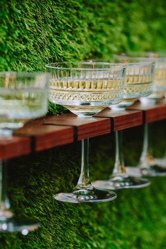 Champagne Glasses And Cocktails Are Mounted On A Stand On The Vertical Wall Of The Green Lawn. Decor For A Wedding Or Party In The Italian Style.