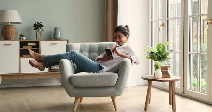 Pretty smiling young Indian woman relax sitting on soft comfy armchair using mobile phone at modern apartment living room interior. Happy millennial female use web application chat online on cellphone