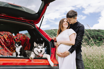 A happy family is expecting a baby and resting with a cat and dog in nature near their car. Travel concept with pets.