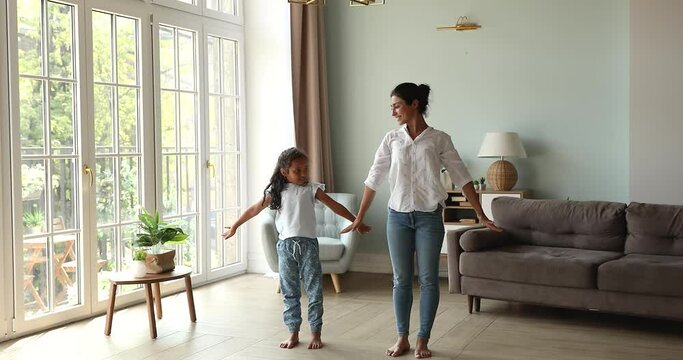 Happy single Indian mother accustom little daughter preschooler to healthy lifestyle teach to do simple yoga exercises. Smiling young mommy yogi enjoy morning gymnastic at living room with child girl